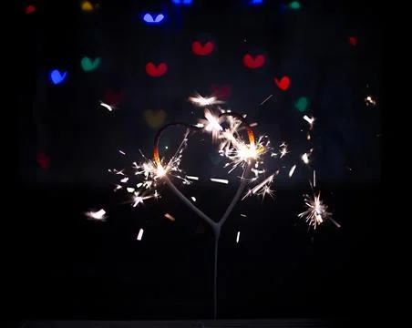 Sparkler in the form of a burning heart surrounded by colorful bokeh Stock Photos