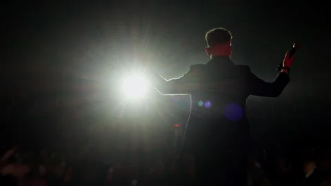 Speaker Gesturing with Open Arms on Stage in Bright Spotlight 動画素材 345100045