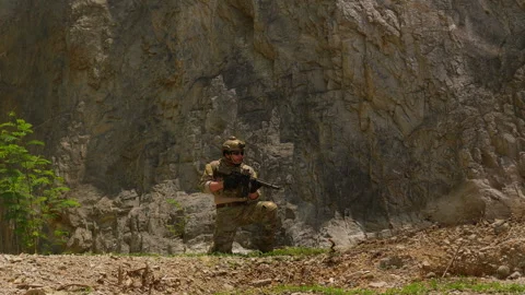 Special forces soldier in combat gear kneels with a rifle at the base of a cliff Stock Footage 342620602