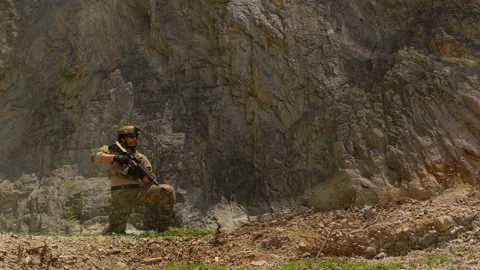 Special forces soldier in combat gear kneels with a rifle at the base of a cliff Stock Footage 342620671