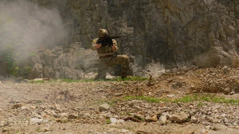 Special forces soldier in combat gear kneels with a rifle at the base of a cliff Stock Footage 342620680