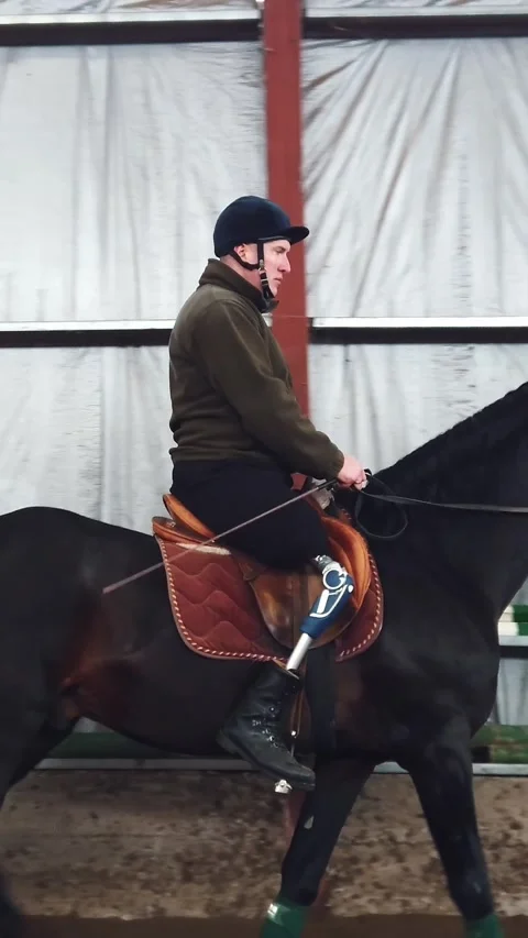 In special hangar, a young disabled man learns to ride a black, thoroughbred Stock Footage 315563487