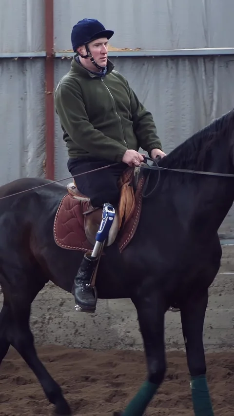 In special hangar, a young disabled man learns to ride a black, Thoroughbred Stock Footage 315757362