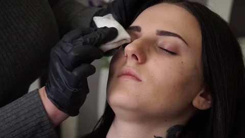 Specialist of permanent make-up wipes the face of the female patient with a Stock Footage 83328425