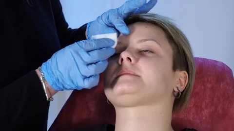 Specialist of permanent make-up wipes the face of the female patient with a Stock Footage 85616345