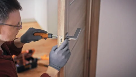 A specialist in work gloves fixes a door handle on a grey brown wooden inter Stock Footage 340786542