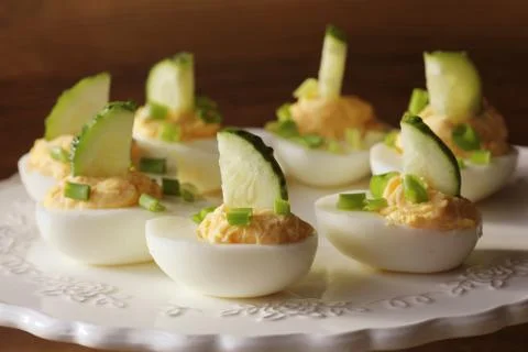Spicy deviled eggs garnished with cucumber and leek on white plate. Wooden ru Stock Photos