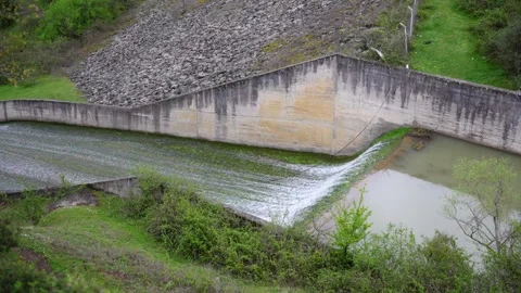 Spillway and dam structure with water flowing over a weir, surrounded by foliage Stock Footage 319791902