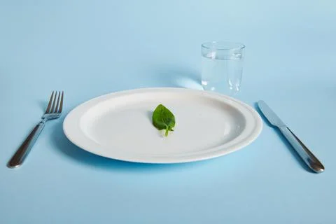 A spinach leaf on a plate, fork, knife and glass with water on blue backgroun Stock Photos