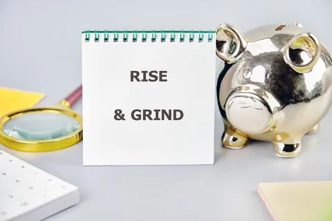 A spiral notepad displaying the words RISE GRIND stands next to a golden Piggy Stock Photos