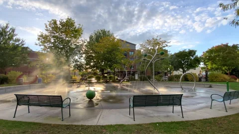 Splash pad water park in a public park with green benches and trees under a cl-1 Stock-Footage 345936604