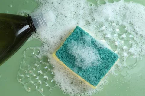 Sponge with foam, detergent and bubbles on green background, flat lay Stock Photos