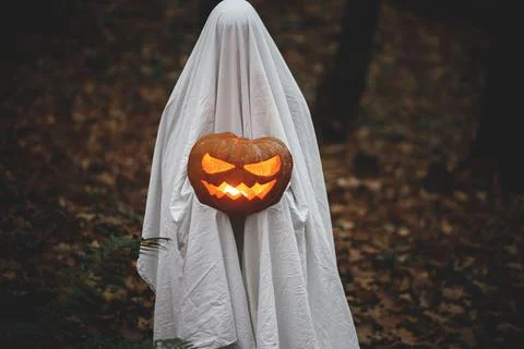 Spooky ghost holding glowing jack o lantern in moody dark autumn forest. Pe.. Stock Photos
