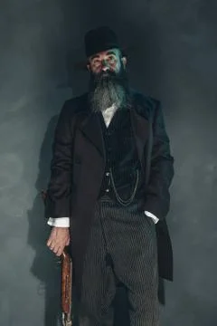 Spooky vintage beard man with rifle in 1900 style fashion against grey wall. Stock Photos