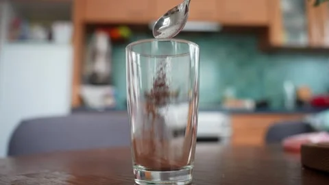 Spoon drops cocoa powder into a tall glass on a wooden table, close-up with Stock Footage 327829919