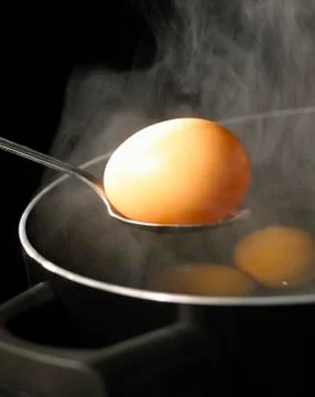 SPOON HOLDING BOILED EGG OVER PAN OF BOILING WATER FULL OF EGGS WITH RISING S Stock Photos