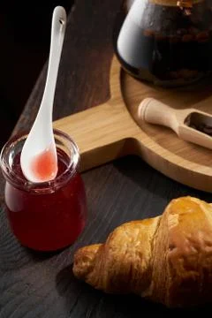 A spoon in a jar of raspberry jam and a croissant on a dark background Board. Stock Photos