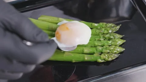 A spoon places a poached egg onto a row of green asparagus spears on a glossy Vídeos de archivo 348688056