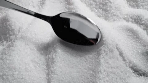 Spoon stirs through white sugar, creating soft waves in surface. Light reflects Vidéo 329436760