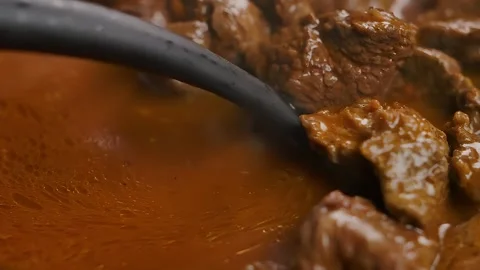 A spoon turns a piece of beef that is stewing in sauce in a frying pan. Close-up Stock Footage 295657105