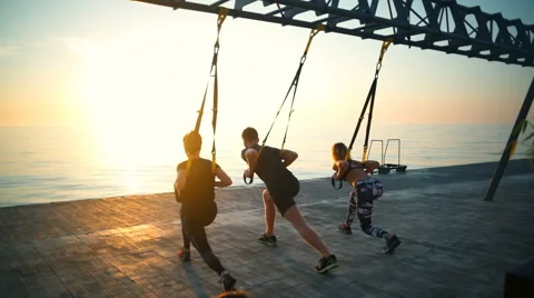 Sportive people training squats with trx at seashore against sun. Slow motion. Stock Footage