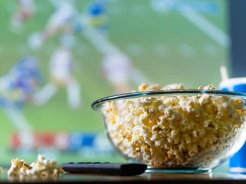A sports program on the TV screen, a large bowl of popcorn and a TV remote co Stock Photos