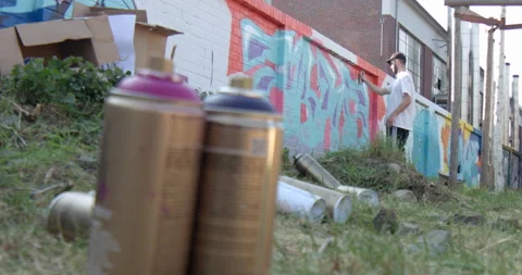 Spray paint cans on the ground and a male graffiti painter drawing on a wall Stock Footage 238857104