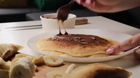 Spreading chocolate paste on pancakes with spoon, served with banana slices on Stock Footage 316472579