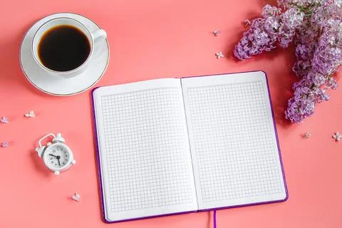 Spring flatlay. Notepad, lilac flowers and cup of coffee on coral background. Stock Photos
