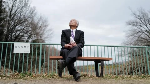 In a spring park with fresh greenery, a man in his 70s wearing a suit sits on a Video stock 307796973