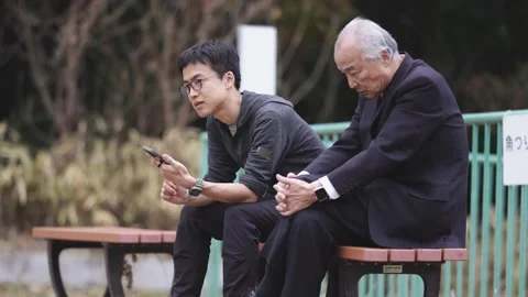 In a spring park, a man in his 70s in a suit and a man in his 30s sit on a bench Video stock 307734998