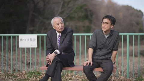 In a spring park, a man in his 70s in a suit and a man in his 30s sit on a bench Stock Footage 307735695