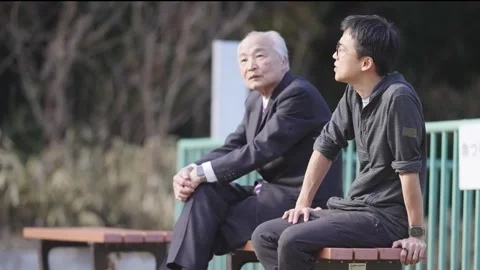 In a spring park, a man in his 70s in a suit and a man in his 30s sit on a bench Stock Footage 307736683