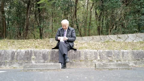 In a spring park, a man in his 70s in a suit and down jacket sits by a parking Stock Footage 307793493