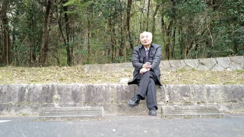 In a spring park, a man in his 70s in a suit and down jacket sits by a parking Vídeo Stock 307793667