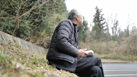 In a spring park, a man in his 70s wearing a suit and down jacket sits by a Stock Footage 307794266