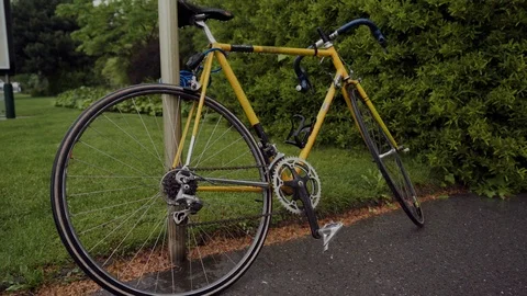 Spring rain falling on yellow city bicycle parked on sidewalk in green park 스톡 동영상 116780339