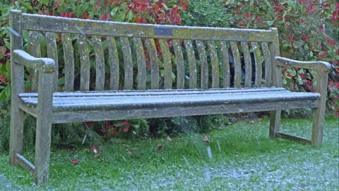 Spring snow falling on an old wooden bench in a garden / yard / park. Video stock 173087665