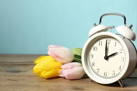 Spring time. Alarm clock and beautiful tulips on wooden table against light.. Stock Photos