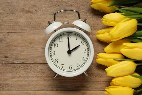 Spring time. Alarm clock and beautiful tulips on wooden table, flat lay Stock Photos