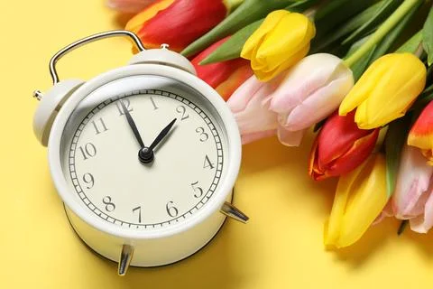 Spring time. Alarm clock and beautiful tulips on yellow background, above vie Stock Photos