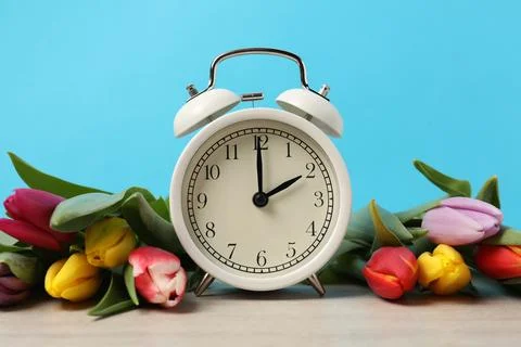 Spring time. Alarm clock and beautiful tulips on wooden table against light.. Stock Photos