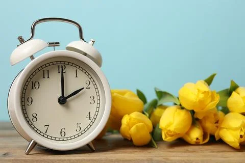 Spring time. Alarm clock and beautiful tulips on wooden table against light.. Stock Photos