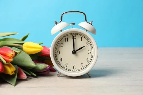 Spring time. Alarm clock and beautiful tulips on wooden table against light.. Stock Photos