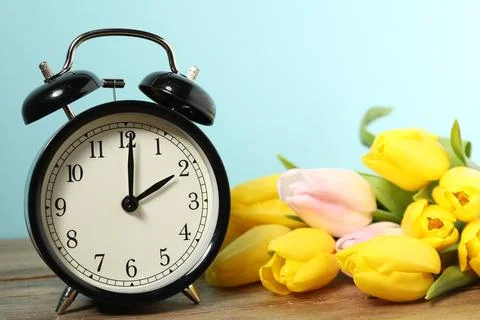 Spring time. Alarm clock and beautiful tulips on wooden table against light.. Stock Photos