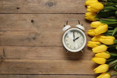 Spring time. Alarm clock and beautiful tulips on wooden table, flat lay. Sp.. Stock Photos
