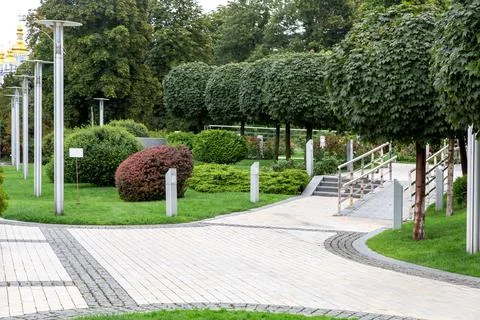 Square paved with stone tiles in a park with green lawns and bushes. Stock Photos