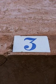 Square white ceramic brick on old wall with the number three painted in blu.. Stock Photos