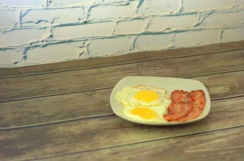 A square white ceramic plate with two fried eggs and slices of bacon stands o Stock Photos
