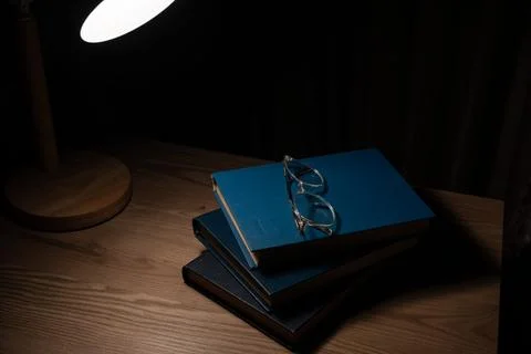 Stack of books with eyeglasses under desk lamp light at night. Stock Photos